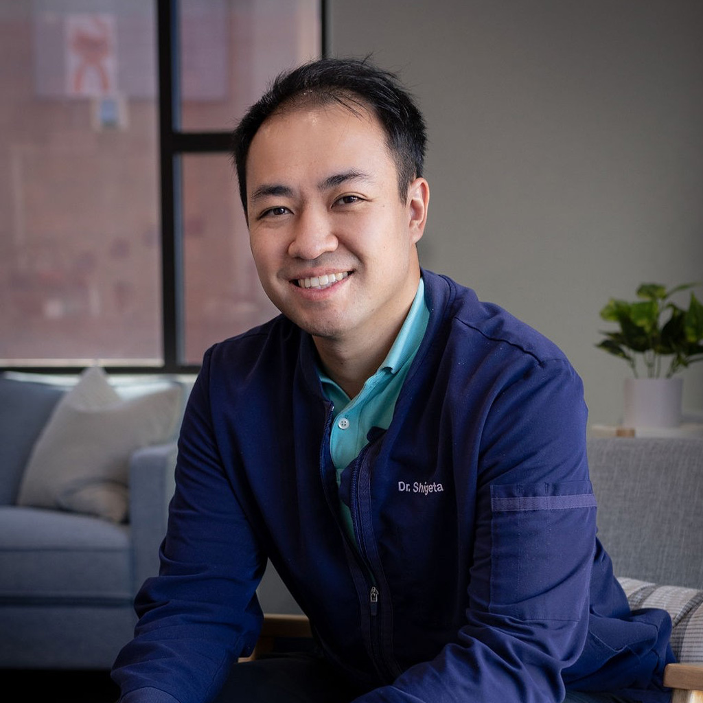 The image features a man sitting on a couch with his legs crossed. He is wearing a blue jacket and has a slight smile on his face, looking directly at the camera.