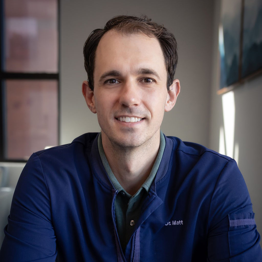 The image depicts a man sitting at a table, smiling slightly, wearing a blue jacket with a name tag, and looking directly at the camera.
