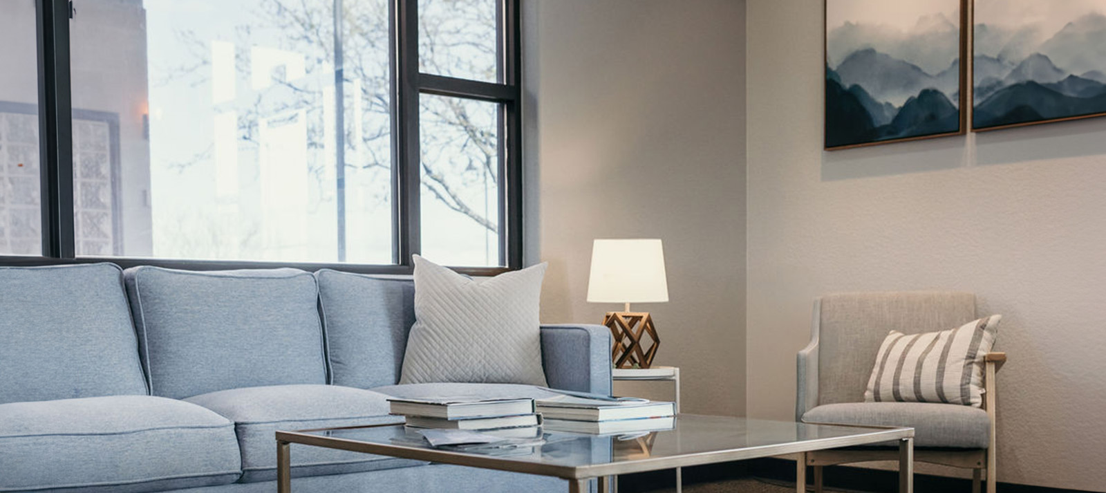 The image shows a modern living room interior with a gray sofa, a coffee table, a rug, a framed picture on the wall, a lamp, and a window allowing natural light into the space.