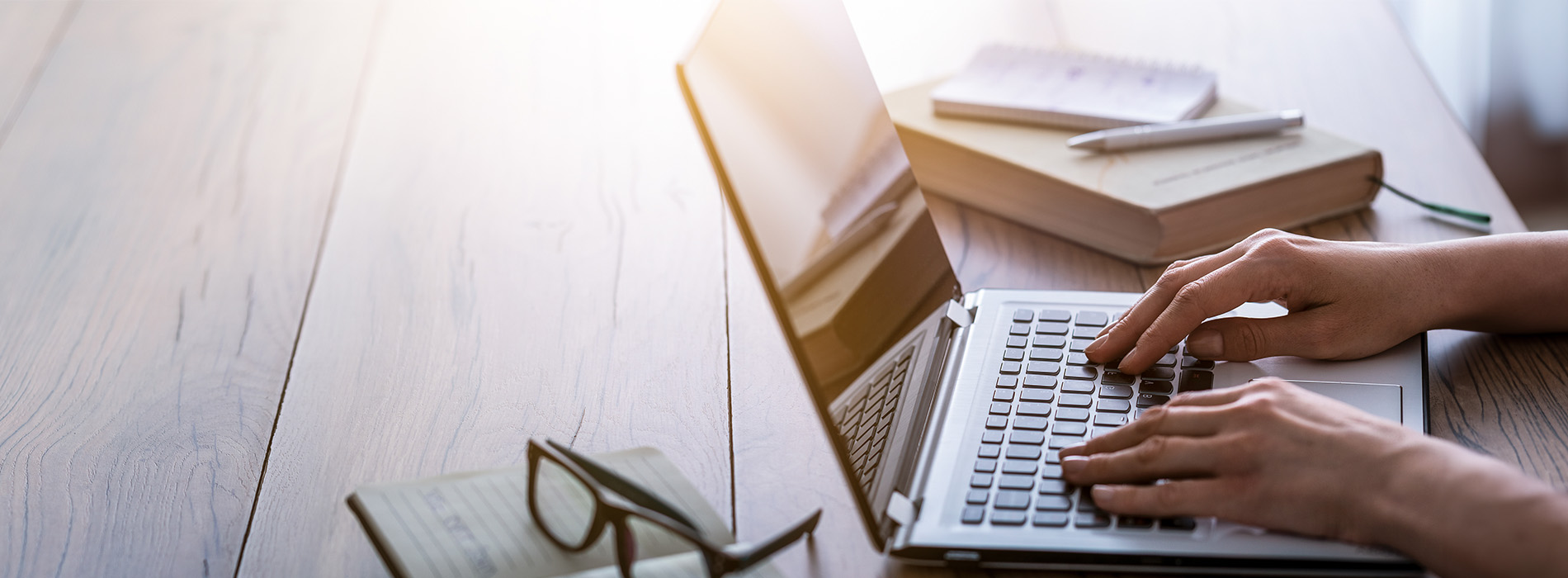 The image features a person typing on a laptop at a desk with various items, including books and papers, set against a blurred background of a room with sunlight filtering through windows.