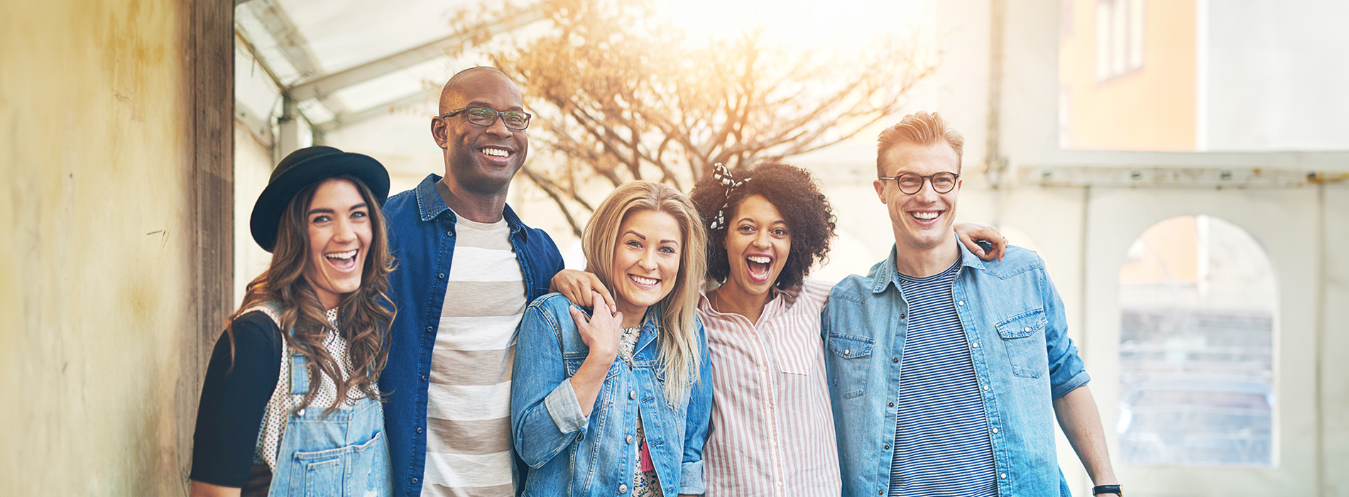 A group of six people posing together, smiling, with some wearing denim jackets, standing outdoors under a building s overhang.