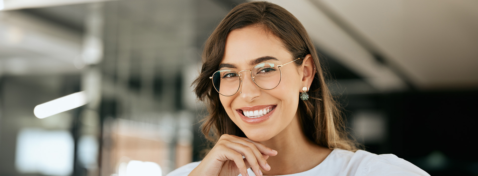 The image features a woman with glasses, smiling at the camera, standing in an indoor setting with a blurred background that suggests a professional environment.