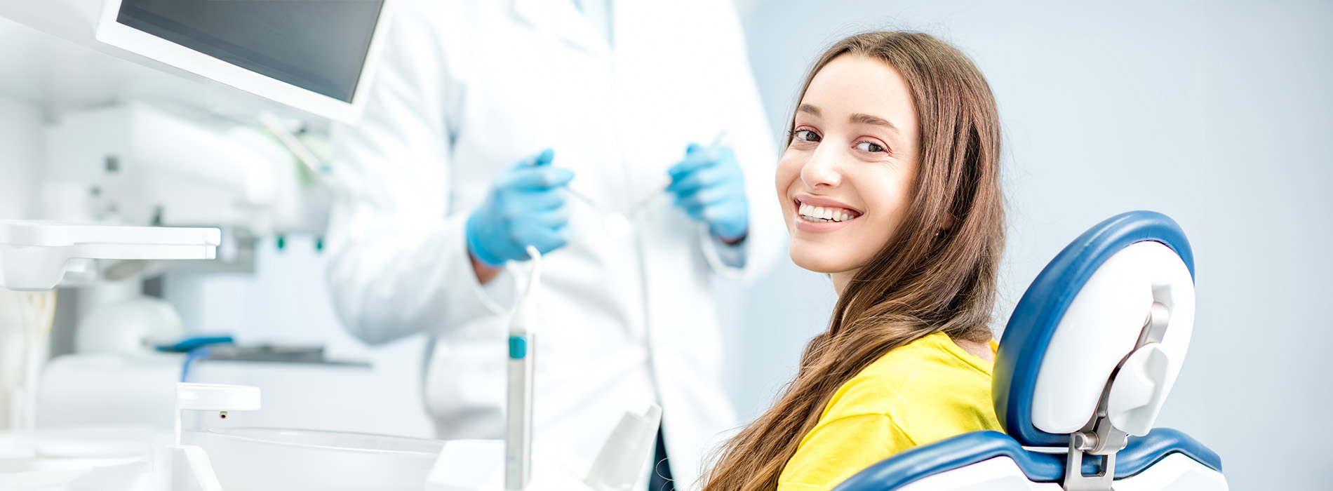 The image shows a woman sitting in a dental chair with a smile on her face, wearing a yellow top, while a dentist stands behind her with a blue gown and stethoscope around his neck in a well-lit dental office setting.