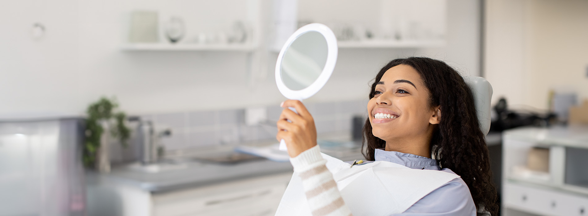 A woman is holding up a mirror while standing in front of a sink with white cabinets in a well-lit room, possibly a bathroom.
