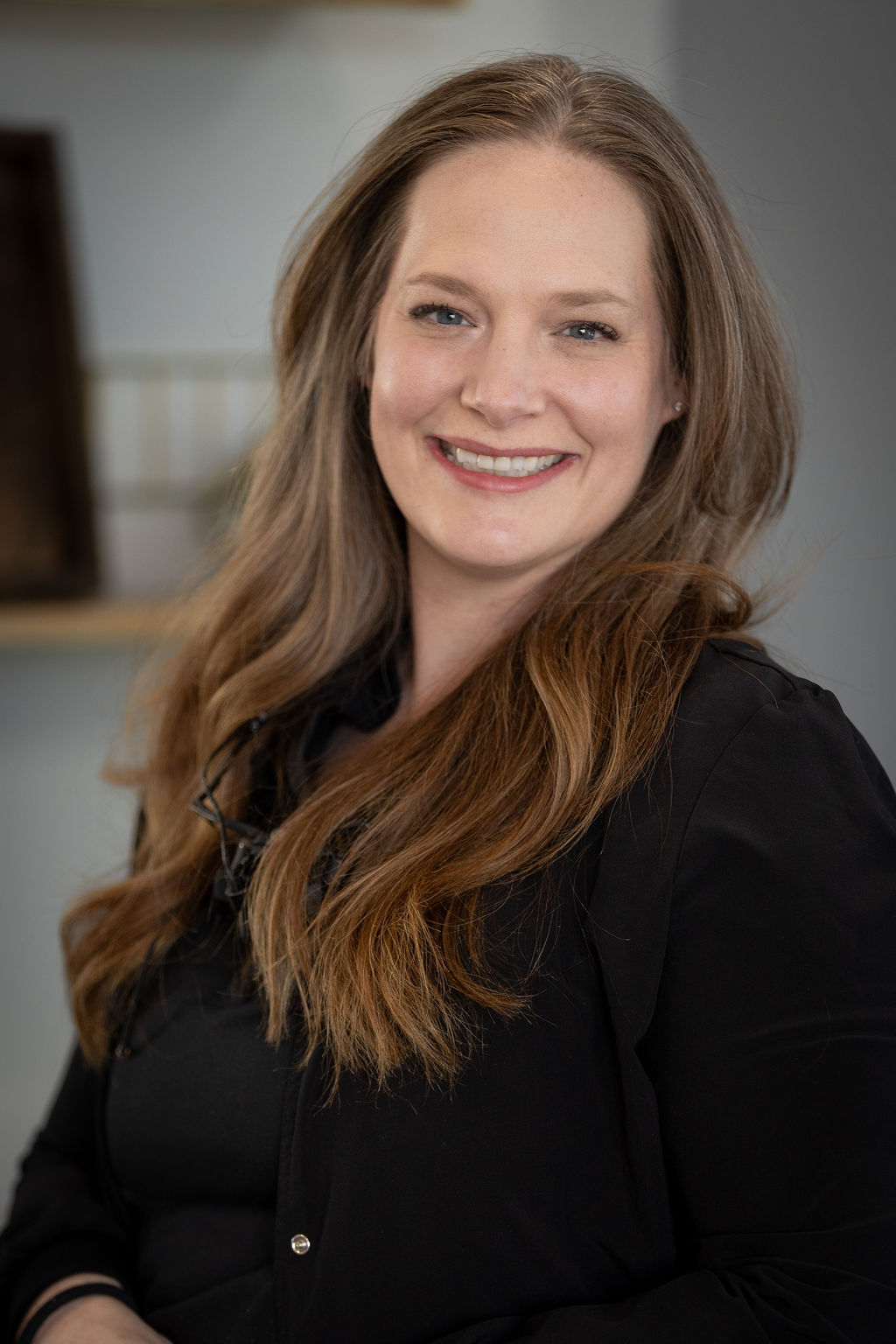 The image shows a woman with long brown hair smiling at the camera. She has a fair complexion and appears to be wearing a black top. Her expression is friendly, and she seems to be posing for the photograph.