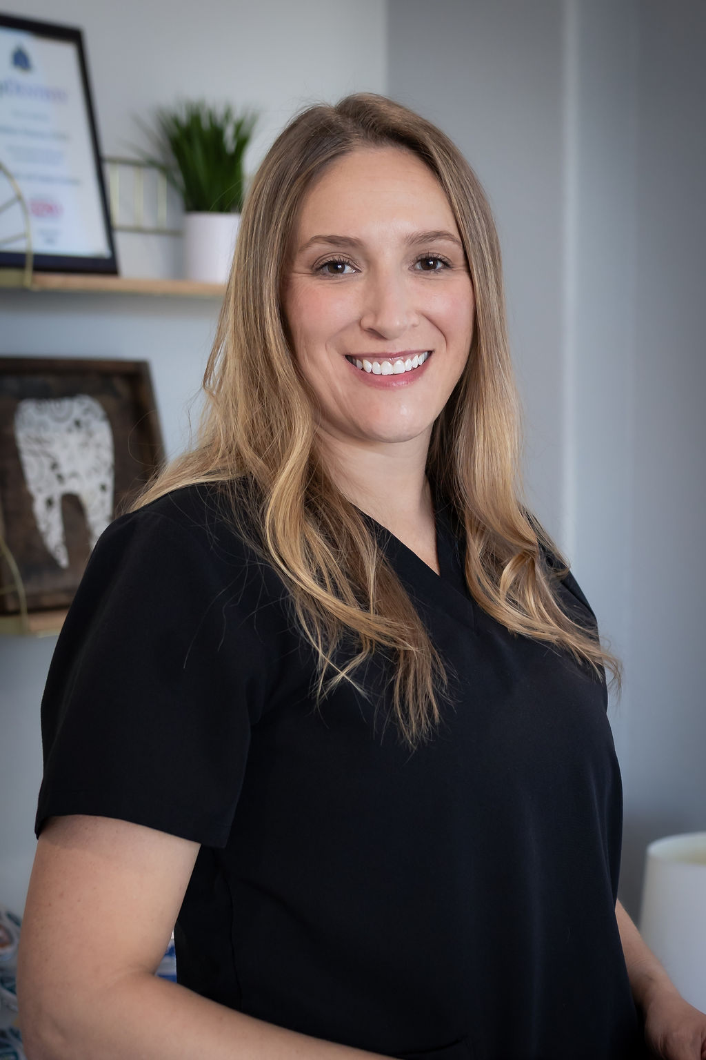 A woman wearing a black shirt stands smiling next to a framed certificate on a wall, with plants and a shelf in the background.
