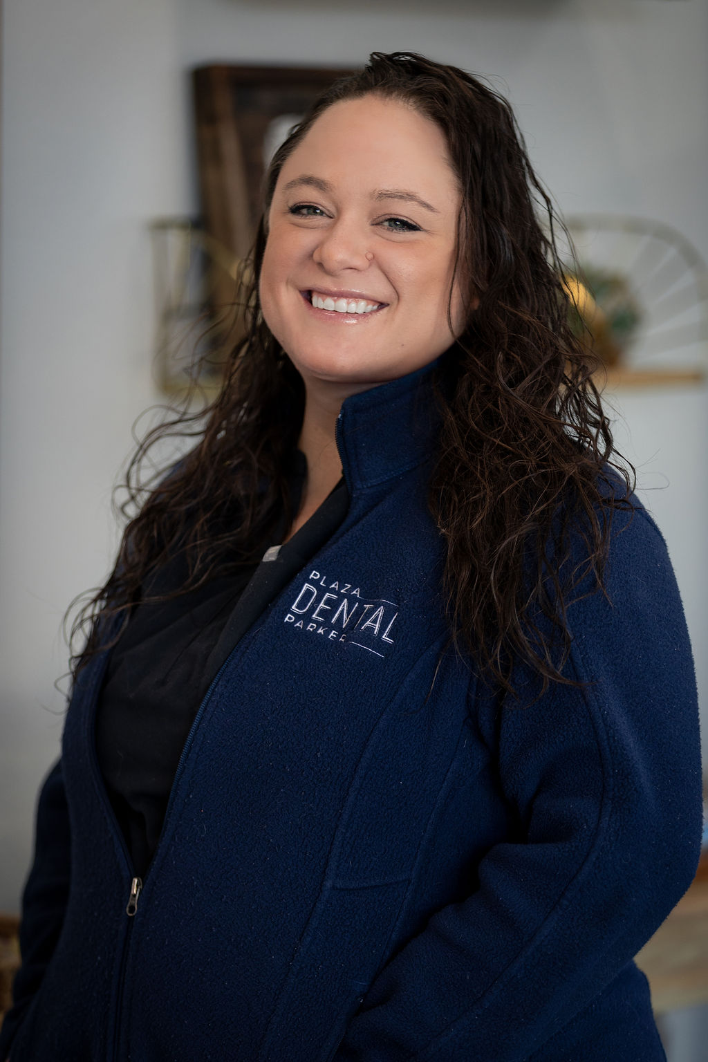The image shows a woman standing indoors, wearing a blue jacket with a name tag on it, smiling at the camera.
