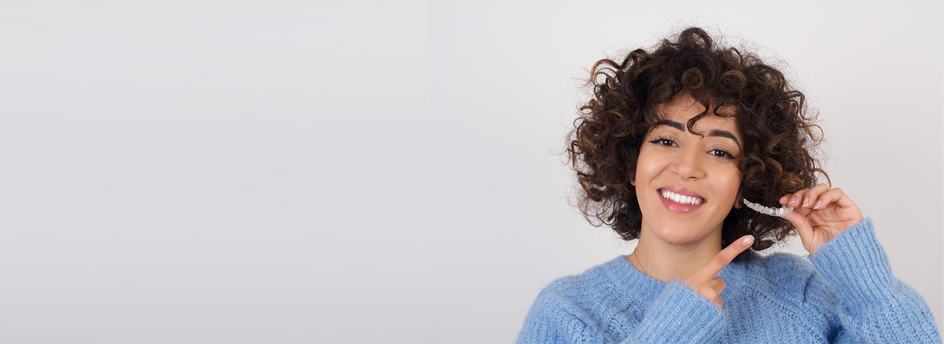 The image shows a person with curly hair smiling at the camera while holding a phone to their ear, wearing a blue sweater, against a white background.