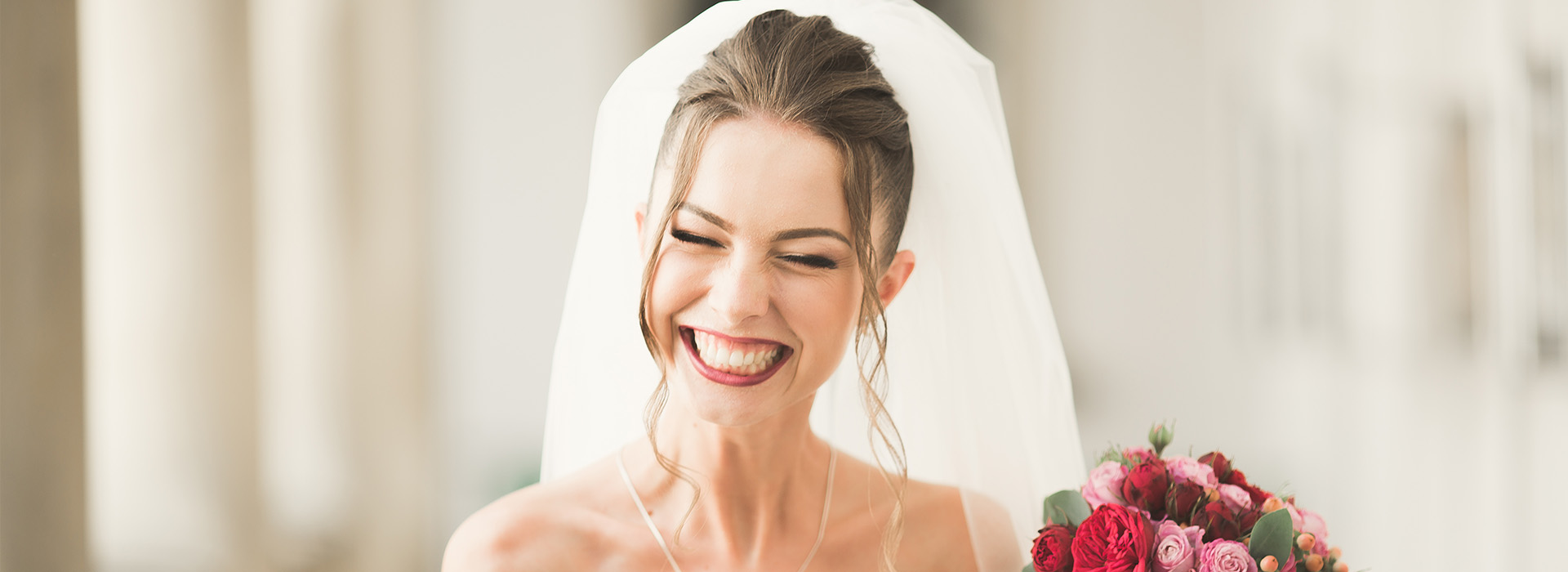The image shows a bride wearing a white wedding dress with a veil, standing next to a bouquet of flowers, smiling, and looking towards the camera.