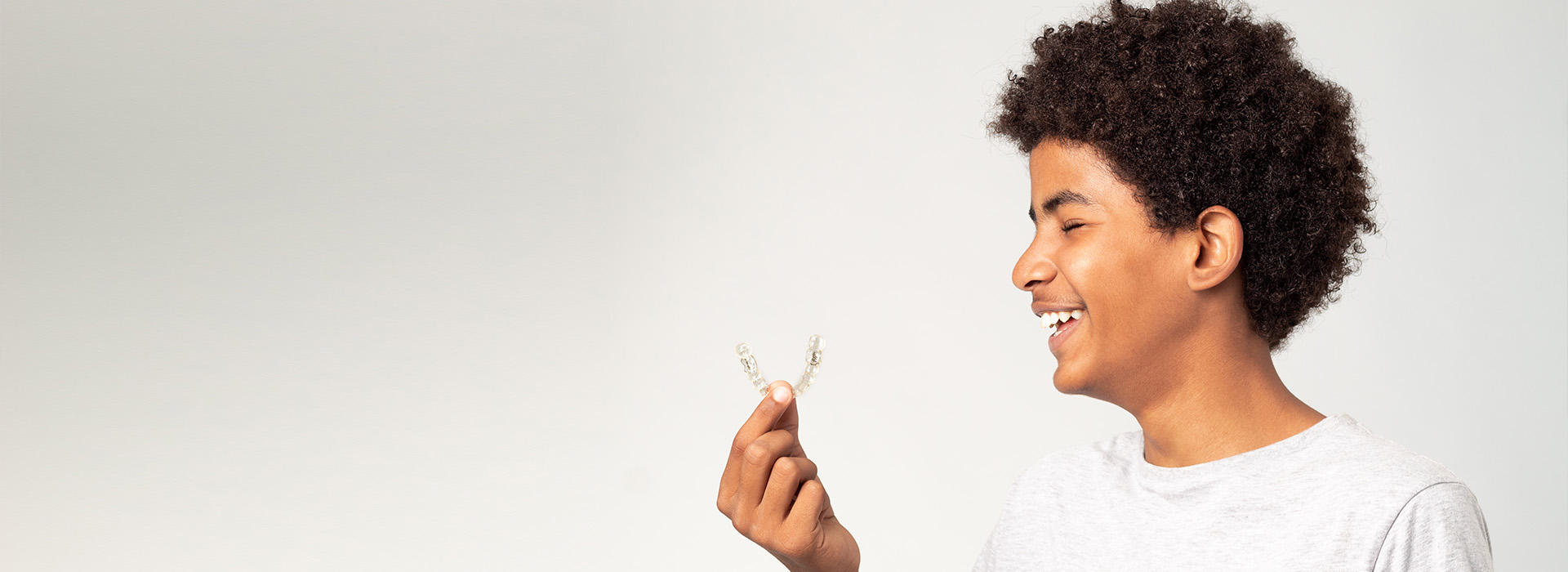 The image shows a young person with dark hair wearing a white top, holding a small object near their face with a smile, against a plain background.