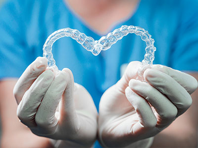 The image shows a person s hands holding a transparent heart-shaped object with a clear, plastic texture, which appears to be a heart-shaped dental retainer or appliance, against a blue background.