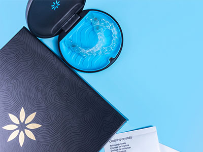 The image shows two open cosmetic product containers with visible contents on a surface against a blue background, accompanied by a closed item that appears to be a packaged product and a small cup of water with a floating object.