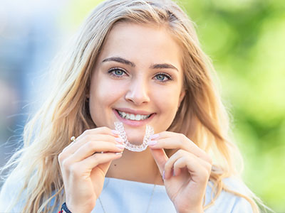 The image shows a young woman holding up a toothbrush with a smile on her face, wearing a necklace, and has blue eyes.