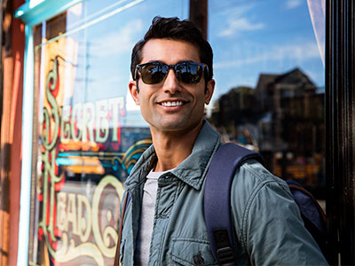 A young man wearing sunglasses and a backpack stands confidently outside a storefront with a sign in the background.