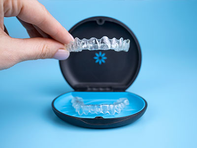 The image shows a person s hand holding an open package of clear aligners, which are transparent orthodontic trays used for teeth straightening, with one tray partially inserted into a blue case that appears to be a mouthguard holder.
