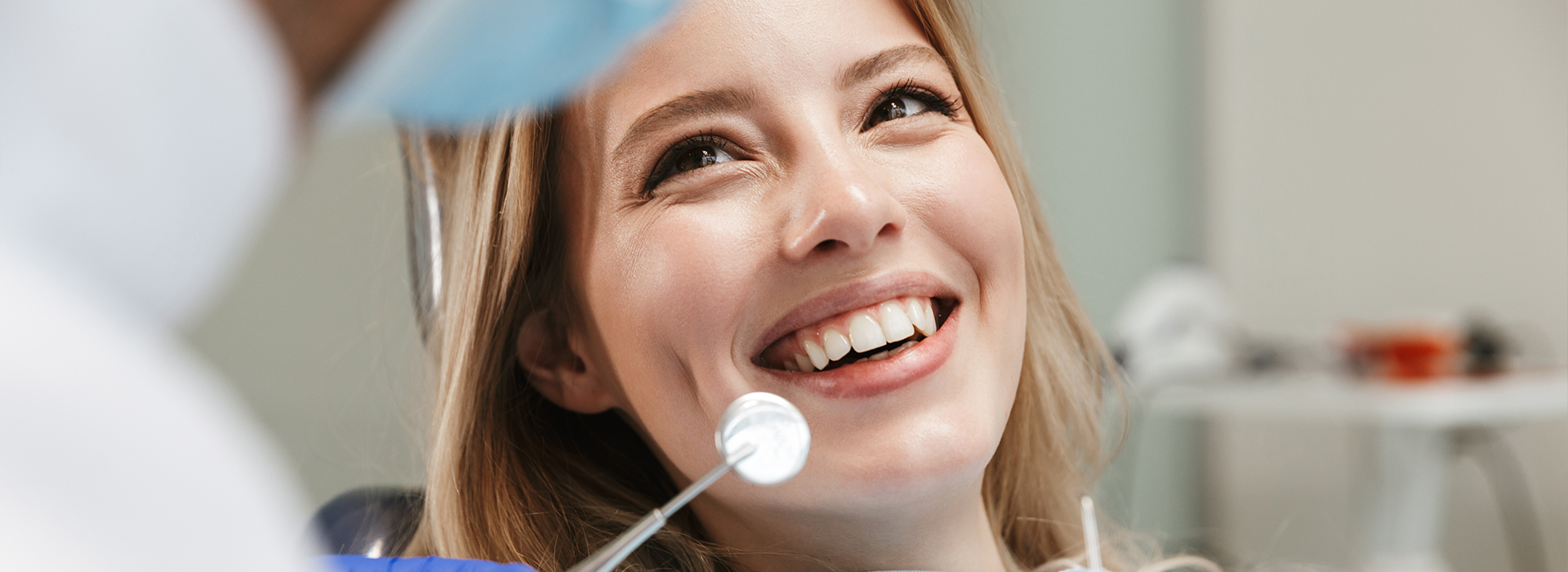 The image shows a woman sitting in a dental chair with a dentist standing beside her, smiling and looking at her with a mirror in hand, likely during a dental appointment.