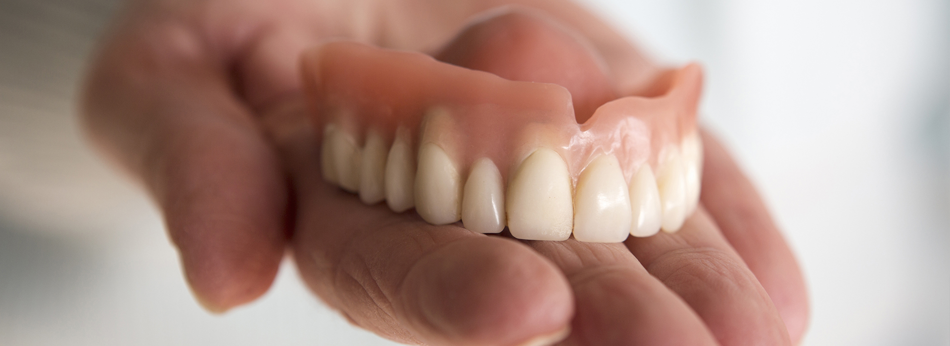 The image shows a person holding a set of dentures with an open mouth, displaying artificial teeth against a blurred background.