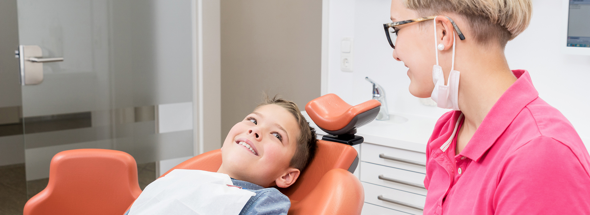 The image shows three individuals in an office setting  a woman seated at a desk with a computer monitor, a young boy sitting in a dental chair receiving dental care from a dentist, and a person standing next to the dentist, likely assisting with the treatment.