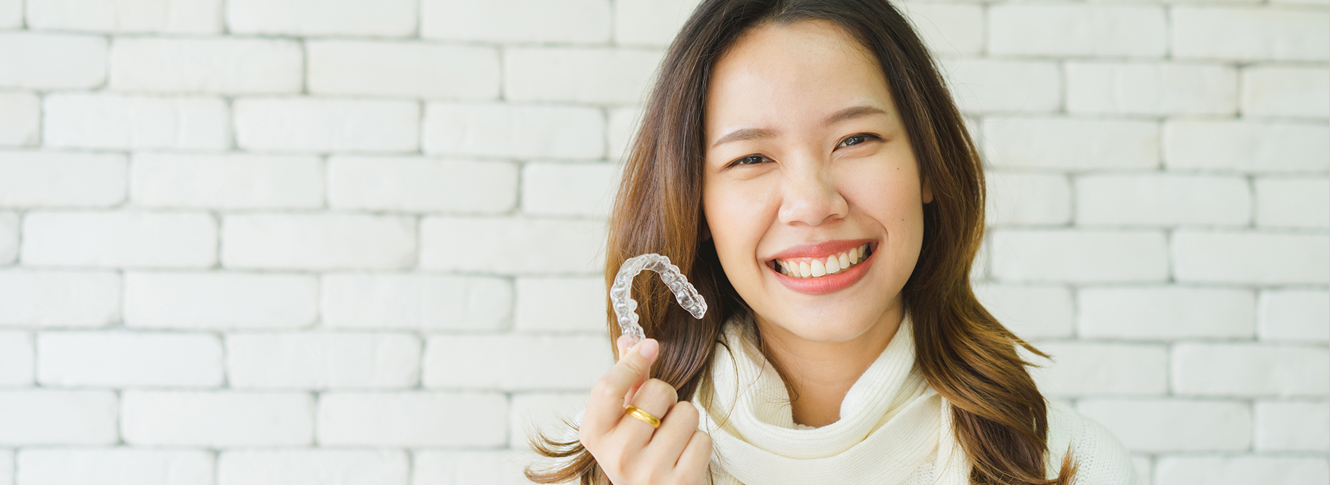 The image shows a smiling woman holding up an object with her right hand against a brick wall background.