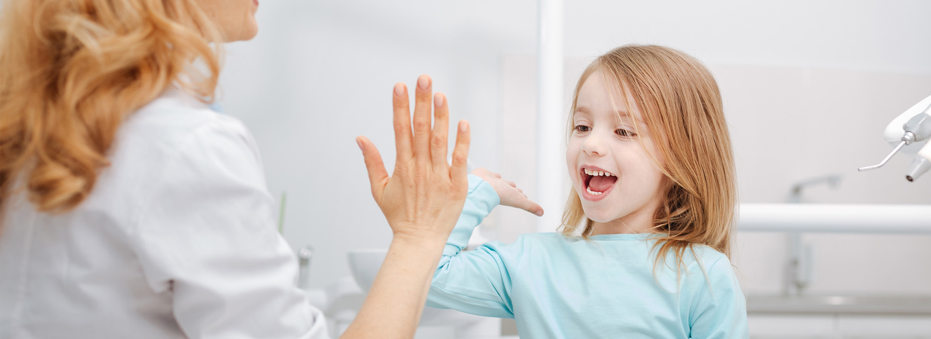 The image shows a young girl with blonde hair looking at her hands clasped together, smiling broadly, while an adult female stands behind her, also smiling and holding up one hand as if waving. Both are in a room that appears to be a bathroom, indicated by the presence of a sink.