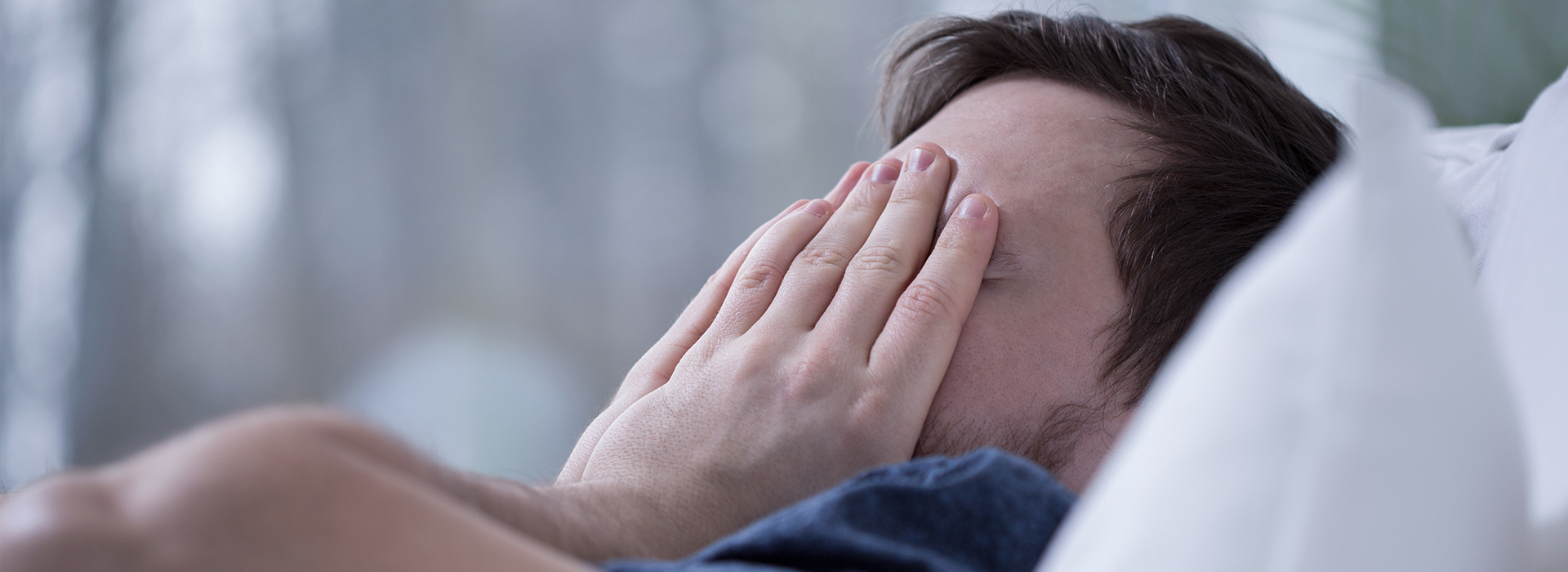 The image shows a man lying in bed with his hand covering his face, appearing to be in a state of distress or discomfort.