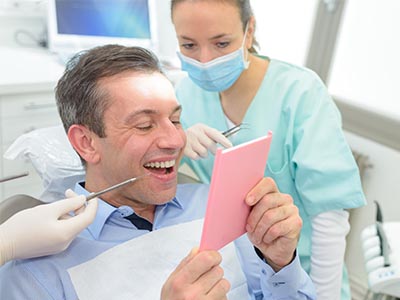A man is sitting in a dental chair with a smile on his face, holding up a pink card, while a dentist and dental hygienist look on.
