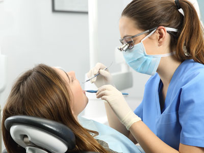 A dental hygienist with a stethoscope around her neck is attending to a woman sitting in a dental chair, performing dental work on her teeth.