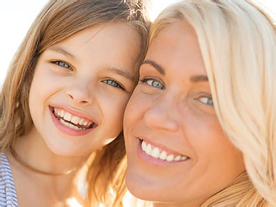 The image shows a woman with a young girl, both smiling at the camera, standing outdoors during daylight.