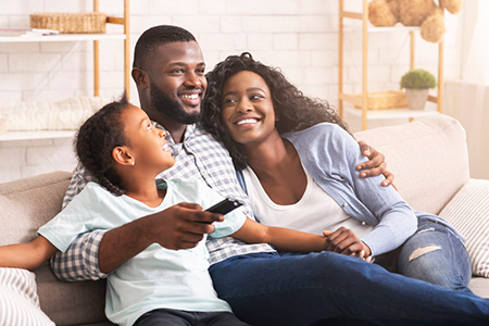 A family of four enjoying a moment together on a couch, with the father holding a remote control and smiling at the camera.