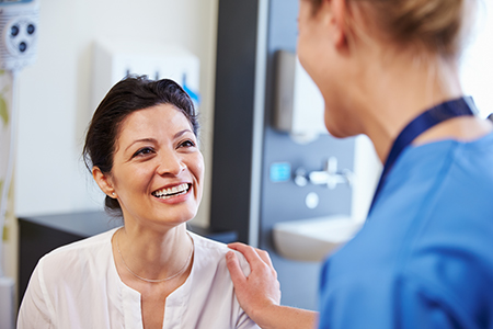 The image shows two individuals in a medical setting  on the left is a person with a smile, wearing a white shirt, and on the right is a woman in a blue uniform, who appears to be a healthcare professional, engaged in conversation with the smiling individual.