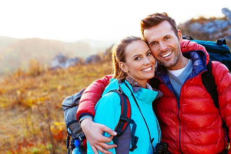 A man and woman posing for a photo outdoors, with both wearing backpacks and standing amidst autumn foliage.