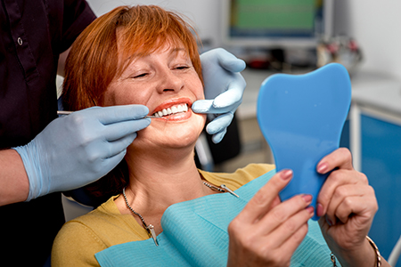 A woman sitting in a dental chair with a blue mouthguard, being attended to by a dentist who is adjusting her teeth, holding up a mirror for her to see her smile.