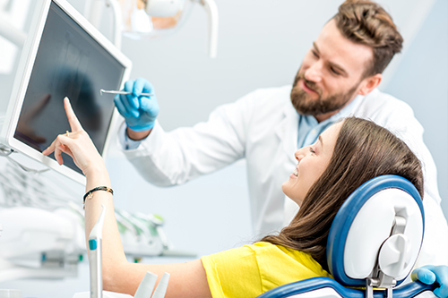 A man and woman in a medical setting, with the man standing and pointing at a computer screen while the woman sits with her head in a dental chair.