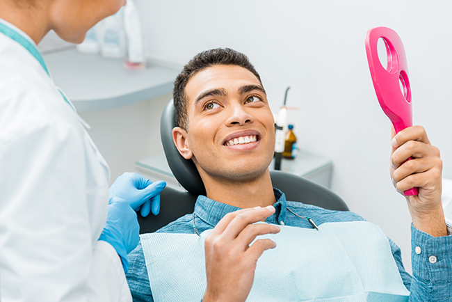 The image shows a man sitting in a dental chair with a smile on his face, holding a pink object in his hand, while a dental professional attends to him.