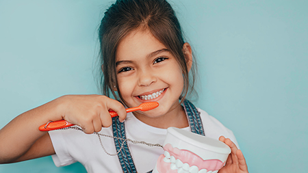 The image shows a young girl brushing her teeth with a red toothbrush while holding a cup of toothpaste.