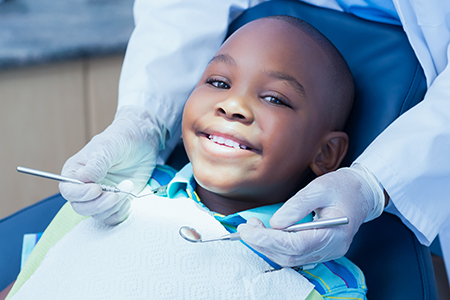 The image shows a young boy sitting in a dental chair, receiving dental treatment while smiling at the camera with a happy expression.