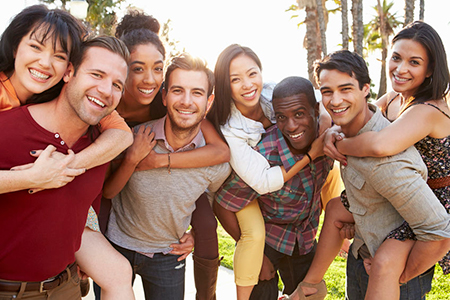 The image shows a group of young adults posing together outdoors, smiling at the camera, with a clear sky in the background.