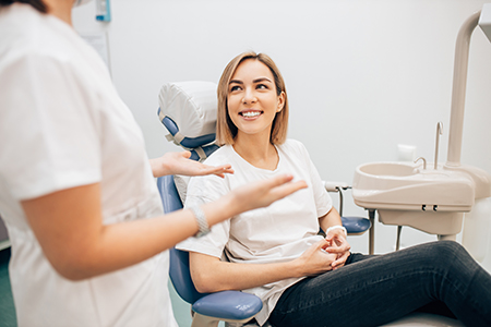 The image shows a woman sitting in a dental chair, smiling at the camera, with a dental professional standing behind her.
