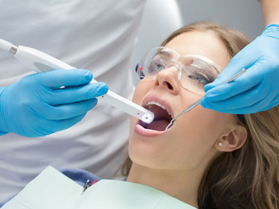 The image shows a woman with her mouth open wide, receiving dental treatment from a dental professional who is using a device to examine her teeth.