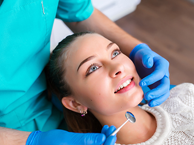 The image shows a woman sitting in a dental chair with a dentist examining her teeth, wearing a blue surgical gown and a stethoscope around her neck.