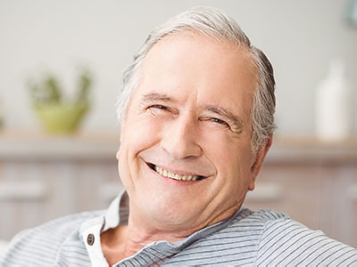 The image shows an elderly man with gray hair smiling while seated in a chair at home.