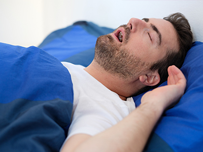A man with facial hair is lying in bed with his eyes closed, appearing to be asleep or resting.