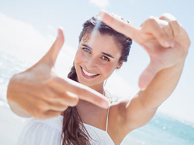 The image shows a young woman with long hair smiling at the camera while holding up her hand to frame a picture of herself. She is wearing a white top and is outdoors, possibly on a beach, given the bright lighting and blue sky background.